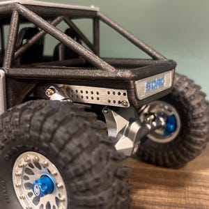 K&ouml;nnte beinhalten: Nahaufnahme eines kleinen, schwarzen und silbernen Offroad-Modellfahrzeugs. Es hat gro&szlig;e, strukturierte Reifen, blaue Akzente und den Text "SDRC". Der Hintergrund ist unscharf, wodurch die Details des Modells hervorgehoben werden.