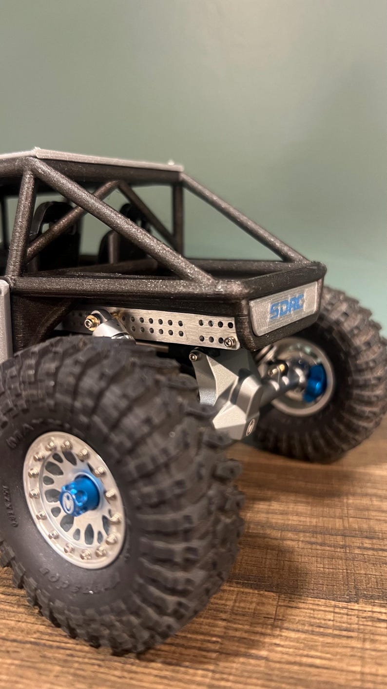K&ouml;nnte beinhalten: Nahaufnahme eines kleinen, schwarzen und silbernen Offroad-Modellfahrzeugs. Es hat gro&szlig;e, strukturierte Reifen, blaue Akzente und den Text "SDRC". Der Hintergrund ist unscharf, wodurch die Details des Modells hervorgehoben werden.