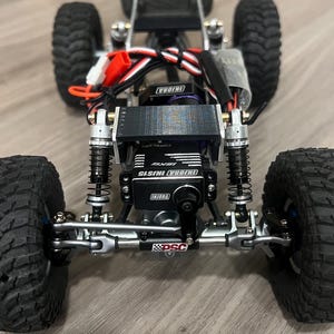 K&ouml;nnte beinhalten: Nahaufnahme eines kleinen, schwarz-silbernen ferngesteuerten Crawler-Trucks. Der Truck hat gro&szlig;e, schwarze, profilierte Reifen, Federung und INJORA-Branding. Das DSC-Logo ist auf der Vorderseite sichtbar.