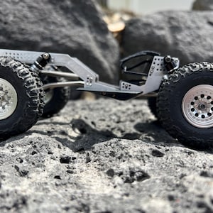 May include: A silver-colored, miniature off-road vehicle with large, black, knobby tires and silver rims. The vehicle is designed with a detailed chassis and suspension system, and is positioned on a rocky surface.
