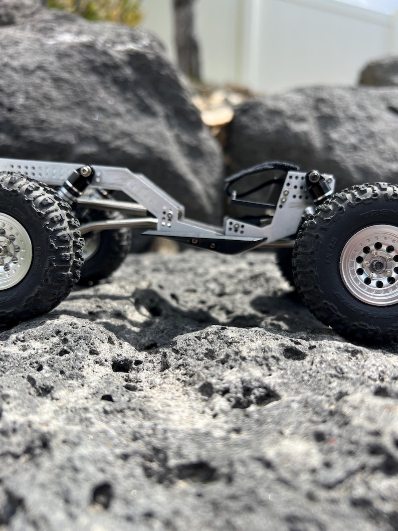 May include: A silver-colored, miniature off-road vehicle with large, black, knobby tires and silver rims. The vehicle is designed with a detailed chassis and suspension system, and is positioned on a rocky surface.