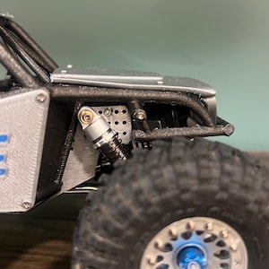 K&ouml;nnte beinhalten: Nahaufnahme eines Miniatur-Gel&auml;ndewagens, der sein robustes Design zeigt. Das Modell verf&uuml;gt &uuml;ber einen schwarzen &Uuml;berrollk&auml;fig, silberne Karosserieteile und gro&szlig;e, strukturierte Reifen. Das Rad hat eine blaue Nabenkappe und silberne Schrauben.