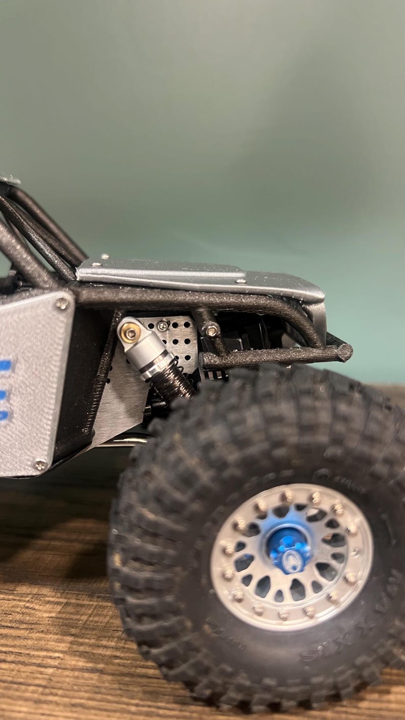 K&ouml;nnte beinhalten: Nahaufnahme eines Miniatur-Gel&auml;ndewagens, der sein robustes Design zeigt. Das Modell verf&uuml;gt &uuml;ber einen schwarzen &Uuml;berrollk&auml;fig, silberne Karosserieteile und gro&szlig;e, strukturierte Reifen. Das Rad hat eine blaue Nabenkappe und silberne Schrauben.