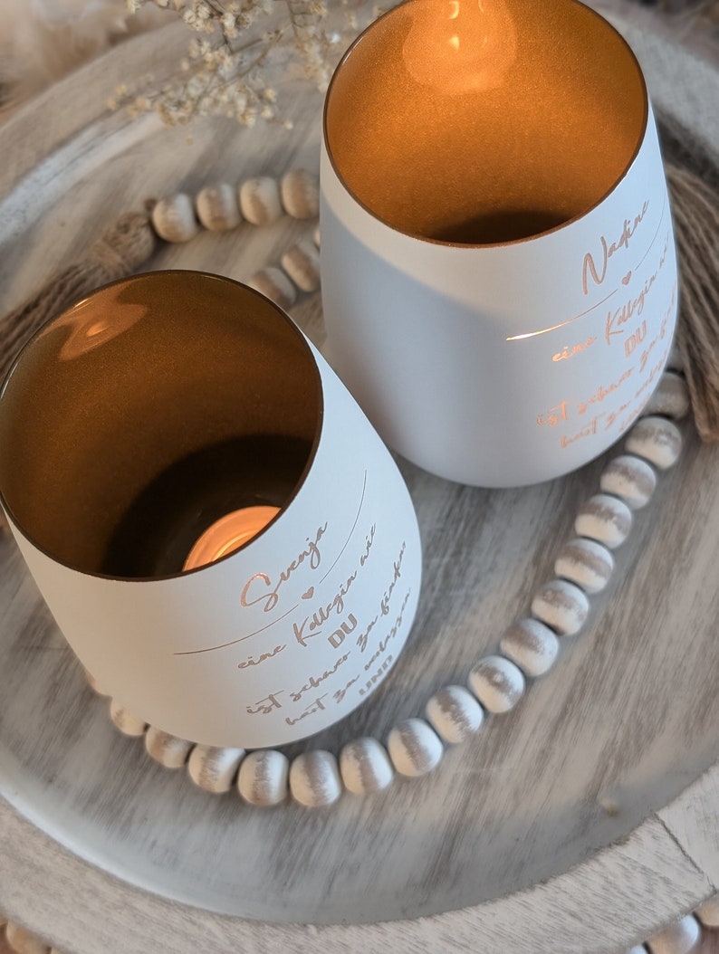 May include: Two white candle holders with gold interiors. The candle holders have personalized messages on them. The message on the left candle holder reads "Svenja, eine Kollegin wie DU ist schwer zu finden,  habt  ihr  ganz  viel  Spa&szlig;  zusammen". The message on the right candle holder reads "Nadine, eine Kollegin wie DU ist schwer zu finden,  habt  ihr  ganz  viel  Spa&szlig;  zusammen".