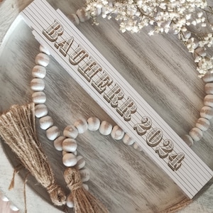 Puede incluir: Una regla de madera blanca con el texto "BAUER 2024" en un estilo vintage tallado. La regla está colocada sobre una bandeja redonda de madera envejecida, rodeada de una guirnalda de cuentas y flores secas. Decoración rústica.