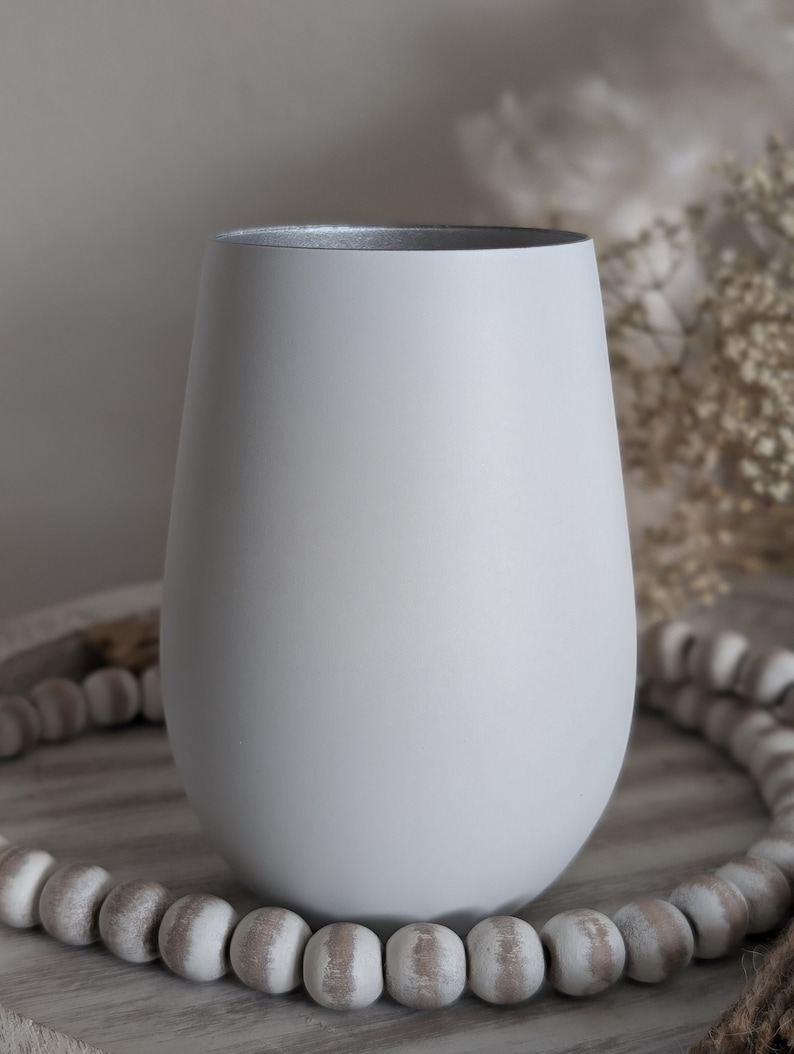 May include: A light gray ceramic vase with a rounded bottom and a wide opening. The vase is sitting on a wooden surface with a string of white beads.