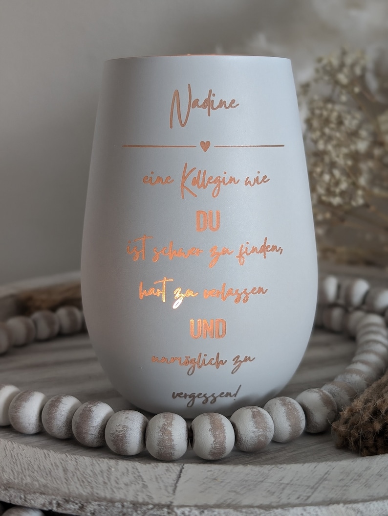 May include: A white ceramic candle holder with a rose gold inscription that reads "Nadine, a colleague like you is hard to find, hard to replace and impossible to forget!"