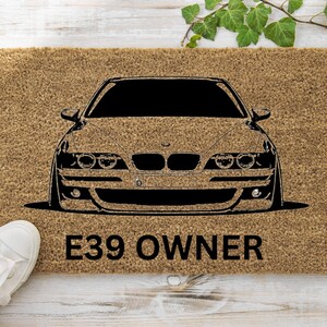 Könnte beinhalten: Eine braune Fußmatte mit einer schwarzen Silhouette eines BMW E39 und dem Text "E39 OWNER".