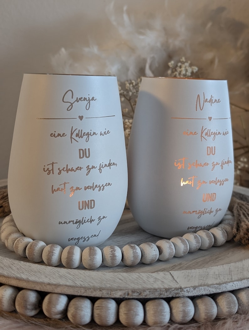 May include: Two white ceramic candle holders with a rose gold metallic finish. The candle holders have a quote about a colleague on each side. The quote on the left says "Svenja, a colleague like you is hard to find, hard to leave and impossible to forget." The quote on the right says "Nadine, a colleague like you is hard to find, hard to leave and impossible to forget."