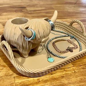 Puede incluir: Una figurita de vaca de las tierras altas de cerámica marrón con una cabeza hueca, utilizada como joyero. La vaca está sentada en una bandeja tejida con una variedad de collares, pulseras y anillos.