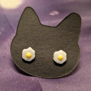 May include: A pair of earrings shaped like fried eggs. The earrings are white with a yellow yolk in the centre. The earrings are displayed on a black cat-shaped card.