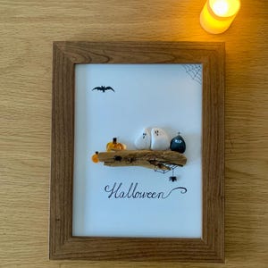 May include: A framed Halloween artwork with a wooden frame. The image shows a bat, spiderweb, and the word "Halloween". It also features a pumpkin, two ghost figures, and a tombstone with "RIP" on it.