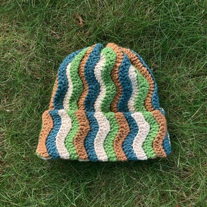 May include: A handmade crochet beanie hat with a wavy pattern in blue, green, brown, and white. The hat is made of soft yarn and is suitable for cold weather. The beanie is shown on green grass. The hat is a stylish accessory.
