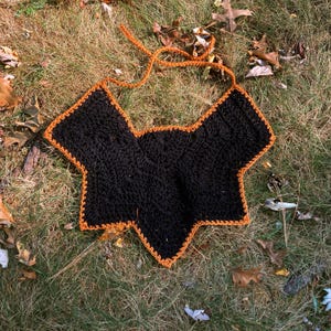 May include: A black crochet top with an orange trim and straps, lying on a bed of grass and fallen leaves. The top has a unique shape and is handmade.