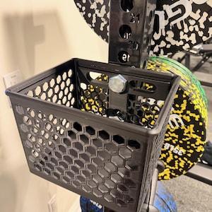 Puede incluir: Una cesta negra para accesorios de rack de levantamiento de pesas con un patrón de panal. La cesta está montada en un rack de metal negro, con discos de pesas en el fondo. Los discos de pesas tienen patrones en blanco y negro y los números 15LB y 35LB.