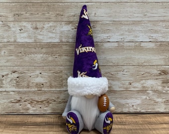 May include: A gnome doll wearing a purple hat with the word "Vikings" and a team logo. The gnome has a white beard, gray body, and is holding a brown football. The gnome's feet also have the team logo.