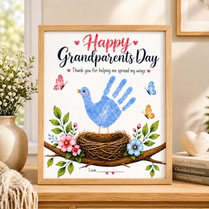 Op de afbeelding: Een ingelijste print met de tekst Happy Grandparents Day en Thank you for helping me spread my wings. Het ontwerp toont een vogel van een blauwe handafdruk in een nest op een tak, omringd door roze en blauwe bloemen en kleurrijke vlinders.