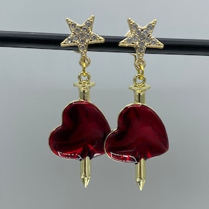 Dangly clip on earrings with red hearts that are pierced by swords, hanging from sparkly stars.