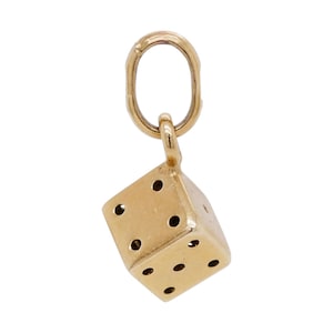 May include: A gold-colored metal charm shaped like a six-sided die, featuring recessed black dots on its faces. The die is attached to a single, oval-shaped jump ring at the top, designed for suspension from a chain or bracelet.