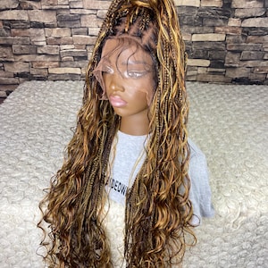 May include: A brown and blonde braided wig with long, curly ends. The wig is styled in a high ponytail with individual braids and a lace front. The wig is displayed on a mannequin head against a brick wall.