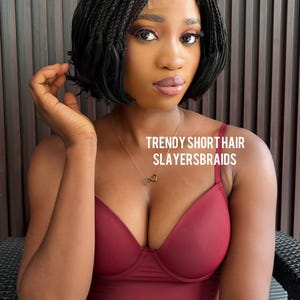 May include: A woman with short, braided black hair and a burgundy top. The text "TRENDY SHORT HAIR SLAYERSBRAIDS" is visible. The woman is wearing a gold necklace and blue jeans.