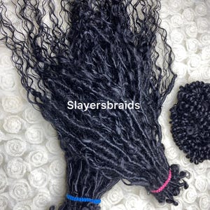 Może przedstawiać: Czarne sztuczne loczki o kręconej fakturze, zebrane i zabezpieczone niebieskimi i różowymi gumkami. Na zdjęciu widoczny jest tekst "Slayersbraids". Loczki ułożone są na białej, teksturowanej powierzchni.