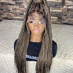 May include: A braided wig with a lace front, displayed on a mannequin head. The wig features a mix of blonde and dark brown braids, styled with some braids pulled up. The mannequin is wearing a black shirt with the word "BRAIDED" printed on it.