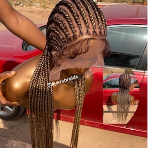 May include: A brown braided wig with long, straight strands. The wig is styled in a cornrow pattern and is worn on a mannequin head. The wig is displayed against a red car.
