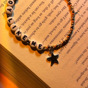 May include: A beaded bracelet with the words "LIKE ME" spelled out in white letter beads. The bracelet has a star charm and is set against a book page.