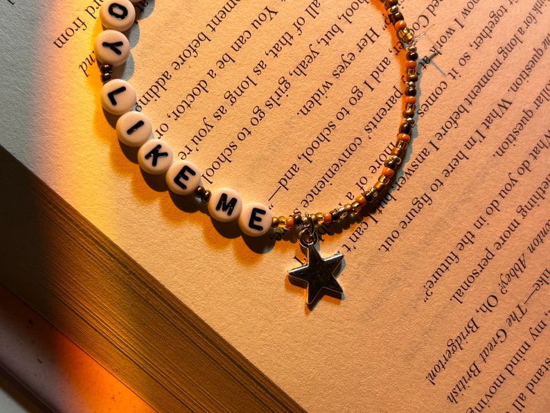 May include: A beaded bracelet with the words "LIKE ME" spelled out in white letter beads. The bracelet has a star charm and is set against a book page.