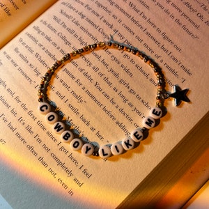May include: A beaded bracelet with white letter beads spelling "COWBOY LIKE ME" is displayed on an open book. The bracelet has gold and black beads, and a star charm. The book's pages are yellowed, and the background is softly lit.