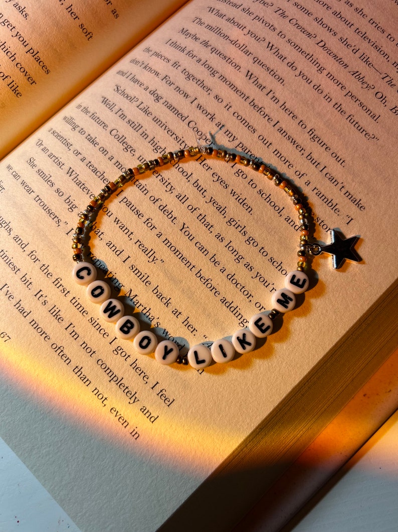 May include: A beaded bracelet with white letter beads spelling "COWBOY LIKE ME" is displayed on an open book. The bracelet has gold and black beads, and a star charm. The book's pages are yellowed, and the background is softly lit.