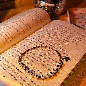 May include: A beaded bracelet with the words "COWBOY LIKE ME" spelled out in white letter beads. The bracelet has gold and brown beads and a star charm. The bracelet rests on an open book.