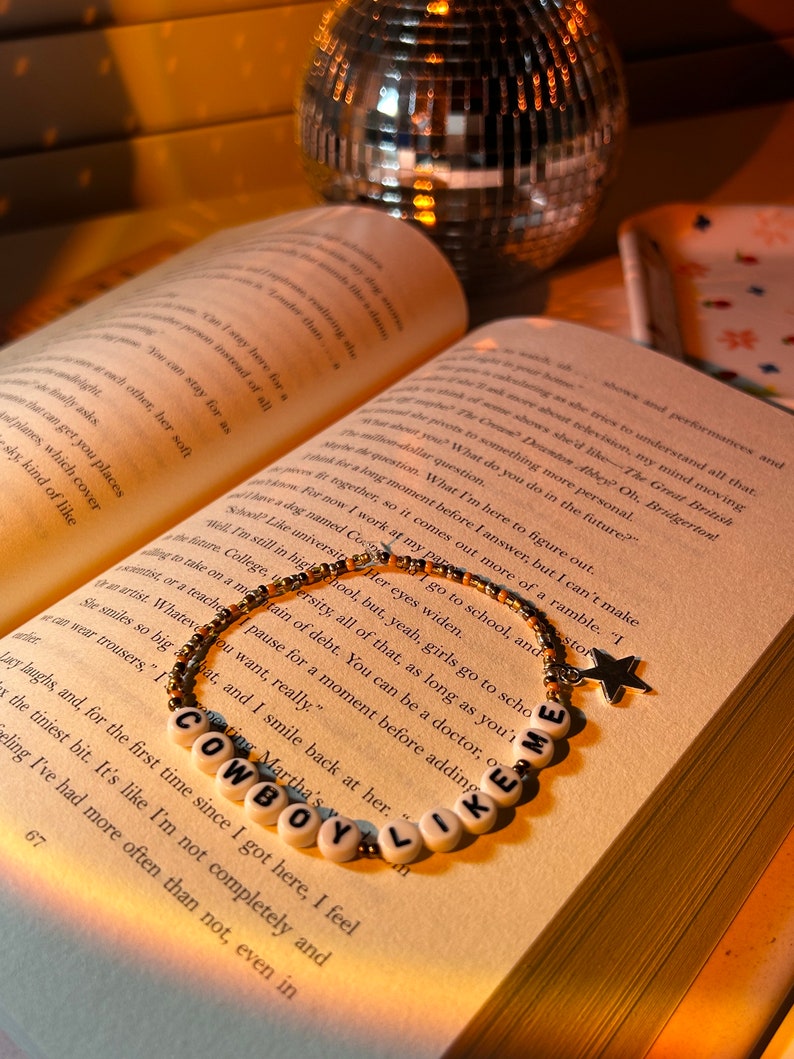 May include: A beaded bracelet with the words "COWBOY LIKE ME" spelled out in white letter beads. The bracelet has gold and brown beads and a star charm. The bracelet rests on an open book.