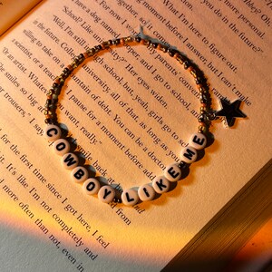 May include: A beaded bracelet with the words "COWBOY LIKE ME" spelled out in white letter beads. The bracelet also features a star charm. The bracelet is resting on the open pages of a book.