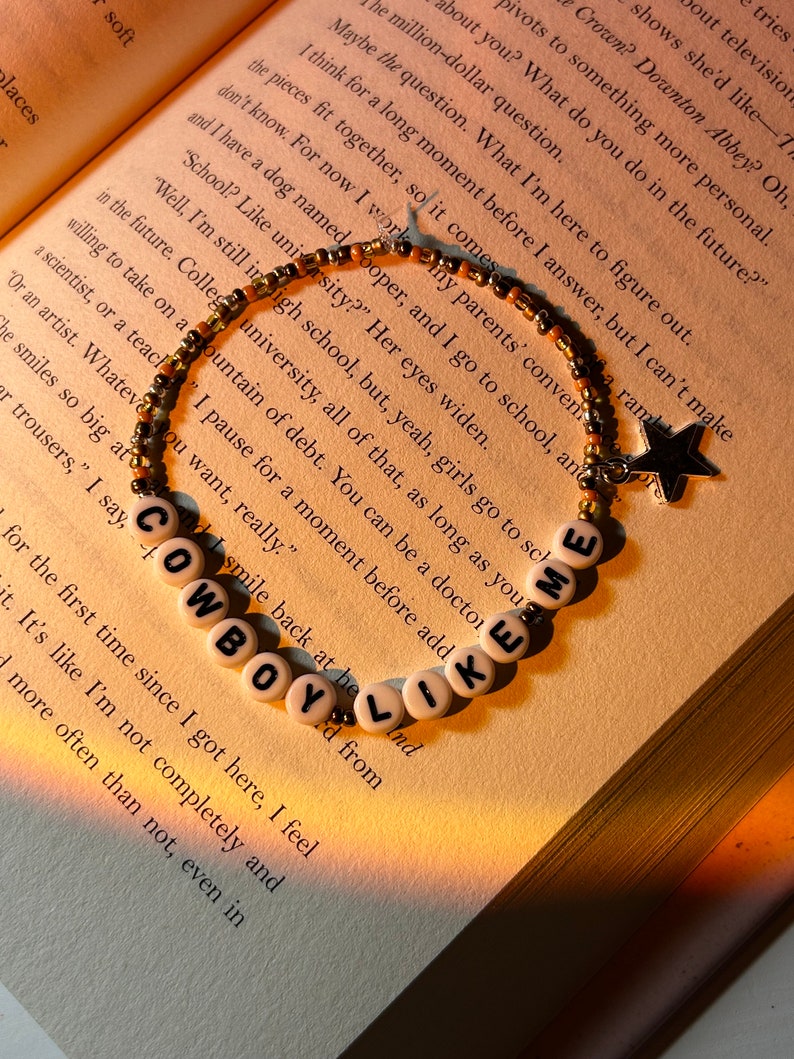 May include: A beaded bracelet with the words "COWBOY LIKE ME" spelled out in white letter beads. The bracelet also features a star charm. The bracelet is resting on the open pages of a book.