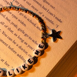 May include: A beaded bracelet with the words "LIKE ME" spelled out in white letter beads. The bracelet has a star charm and is set against a backdrop of a book.