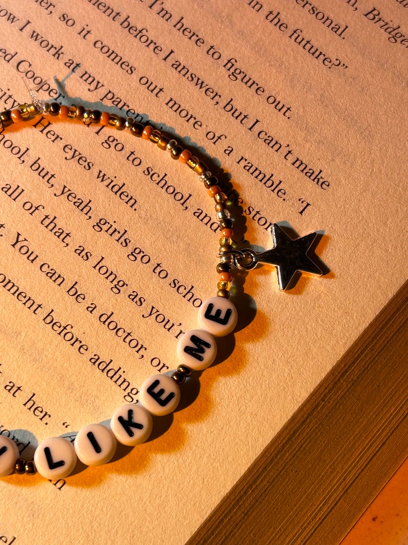 May include: A beaded bracelet with the words "LIKE ME" spelled out in white letter beads. The bracelet has a star charm and is set against a backdrop of a book.