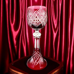 Puede incluir: Una copa de vino de cristal rojo rubí con intrincados diseños grabados. La copa tiene un tallo largo y una base ancha, colocada sobre una superficie de madera oscura contra un telón de fondo de satén rojo. La copa está llena de líquido rojo.