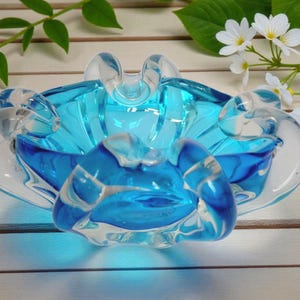 May include: A clear glass ashtray with a vibrant blue interior and clear, curved edges. The ashtray has a unique, organic shape and is set against a light wooden surface, with white flowers and green leaves in the background.