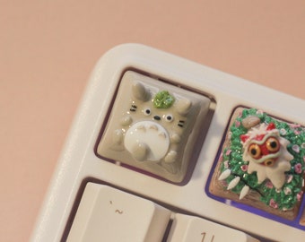 Puede incluir: Un teclado blanco con teclas personalizadas que presentan un personaje Totoro gris, una criatura roja y blanca similar a un león y un plato de cerámica marrón y blanco con un diseño floral.