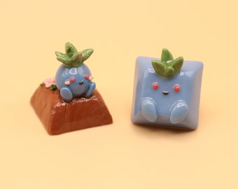 Puede incluir: Dos teclas de Pokemon azules y verdes. Una tecla está sentada sobre una base marrón con flores rosas. La otra tecla es un cuadrado con bordes redondeados.
