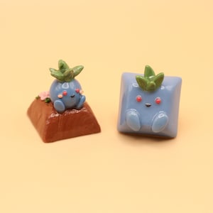 May include: Two blue and green Pokemon keycaps. One keycap is sitting on a brown base with pink flowers. The other keycap is a square with rounded edges.