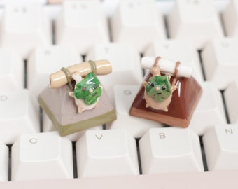 Puede incluir: Dos pequeñas figuritas de cerámica de animales que llevan objetos a la espalda. Las figuritas están sentadas en un teclado blanco. La figurita de la izquierda es verde y marrón y lleva un palo marrón. La figurita de la derecha es marrón y lleva un palo blanco.