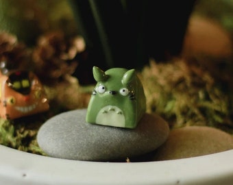 Puede incluir: Una pequeña figurita de cerámica verde de un personaje de dibujos animados con ojos grandes y cuerpo redondeado, sentada sobre una piedra gris en una maceta blanca con musgo verde y otras pequeñas figuritas.