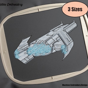 Puede incluir: Diseño de bordado a máquina de una nave espacial gris con detalles azules. El diseño se muestra en una tela gris en un aro. El texto "3 tamaños" se muestra en un óvalo de color melocotón.