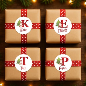 Puede incluir: Cuatro regalos envueltos con cinta roja de lunares y pegatinas personalizadas. Cada regalo tiene una inicial y un nombre diferentes: K para Kaia, E para Elliott, T para Tate y P para Piper. Las pegatinas presentan un árbol de Navidad y Papá Noel.