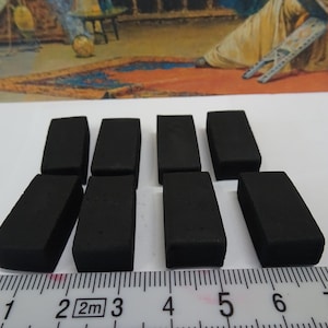 Puede incluir: Una colección de nueve gomas de borrar rectangulares negras dispuestas sobre una superficie blanca. Una regla con marcas de centímetros se coloca debajo. El fondo presenta una pintura con tonos cálidos.