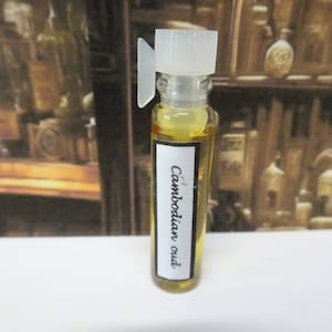 Może przedstawiać: Mała szklana butelka z przezroczystym płynem w środku. Butelka ma białą etykietę z nadrukiem "Cambodian oud".