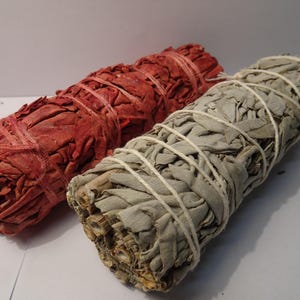 Könnte beinhalten: Zwei Salbei-Bündel, eines rot und eines grau, werden gezeigt. Der Salbei ist mit Bindfaden gebunden. Der rote Salbei hat eine leuchtende Farbe, während der graue Salbei einen gedämpfteren Ton hat. Die Bündel werden wahrscheinlich zum Räuchern verwendet.